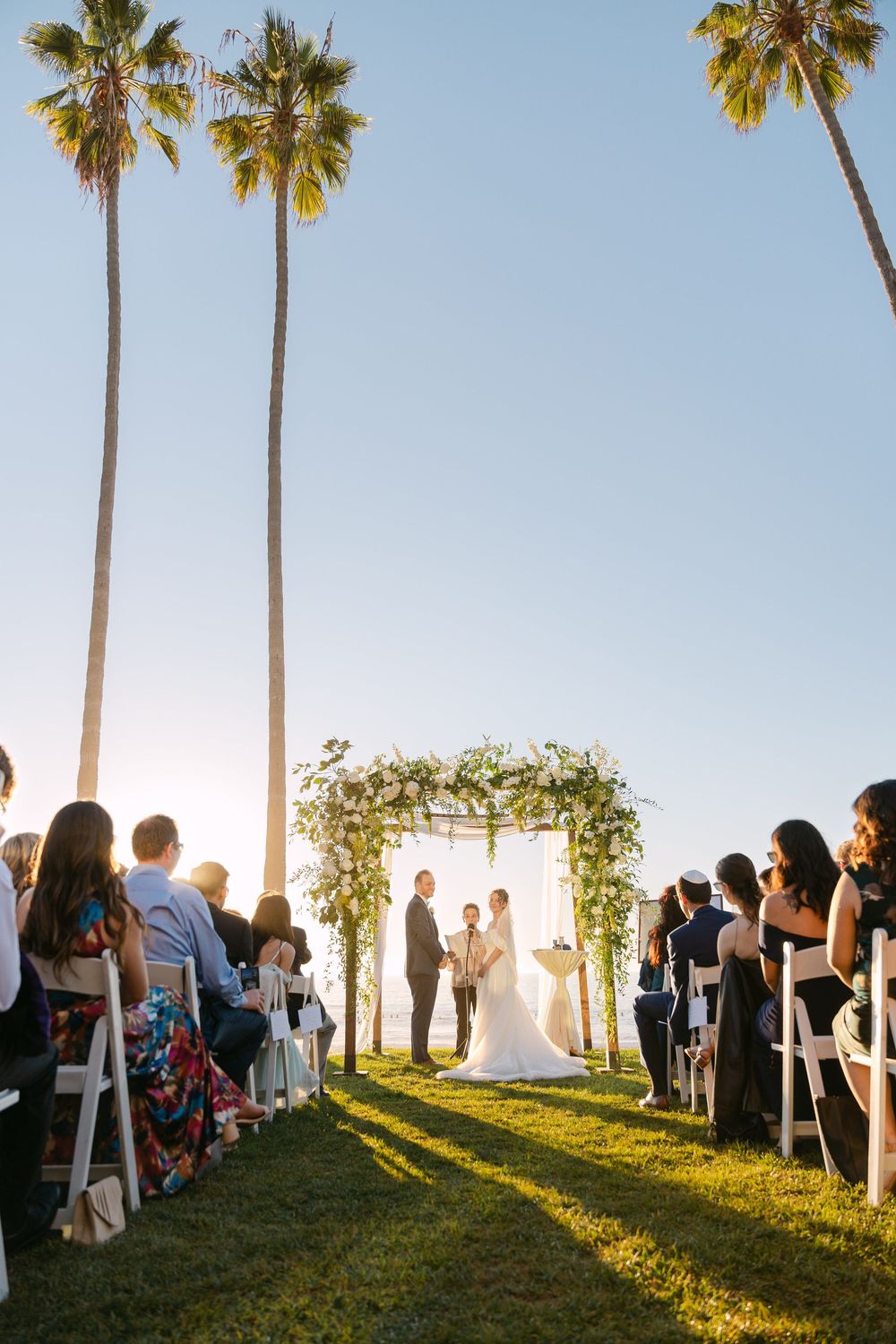 Wedding ceremony guests seated on white chairs under tall palm trees during golden hour sunset.