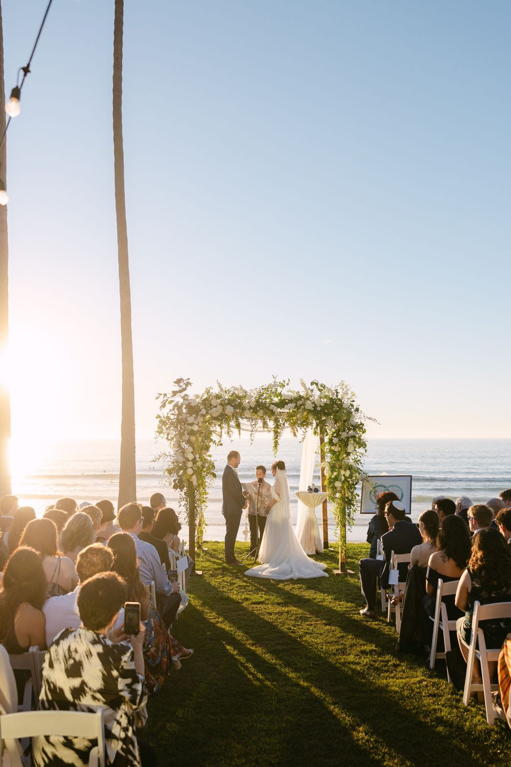 Outdoor wedding ceremony setup on a grassy lawn overlooking the ocean at sunset with palm trees framing the scene.