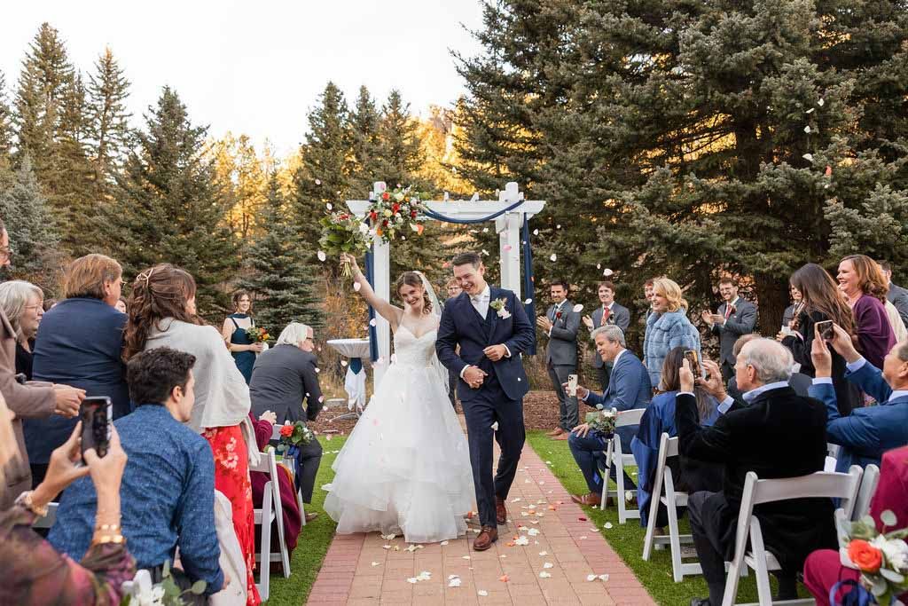 Scenic outdoor wedding ceremony setup with white chairs and wooden arch against mountain and forest backdrop.