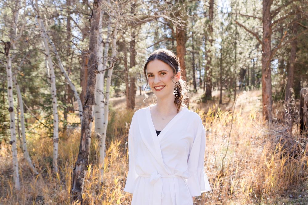 Woman in white button-down shirt poses in autumn forest setting with golden sunlight filtering through trees.