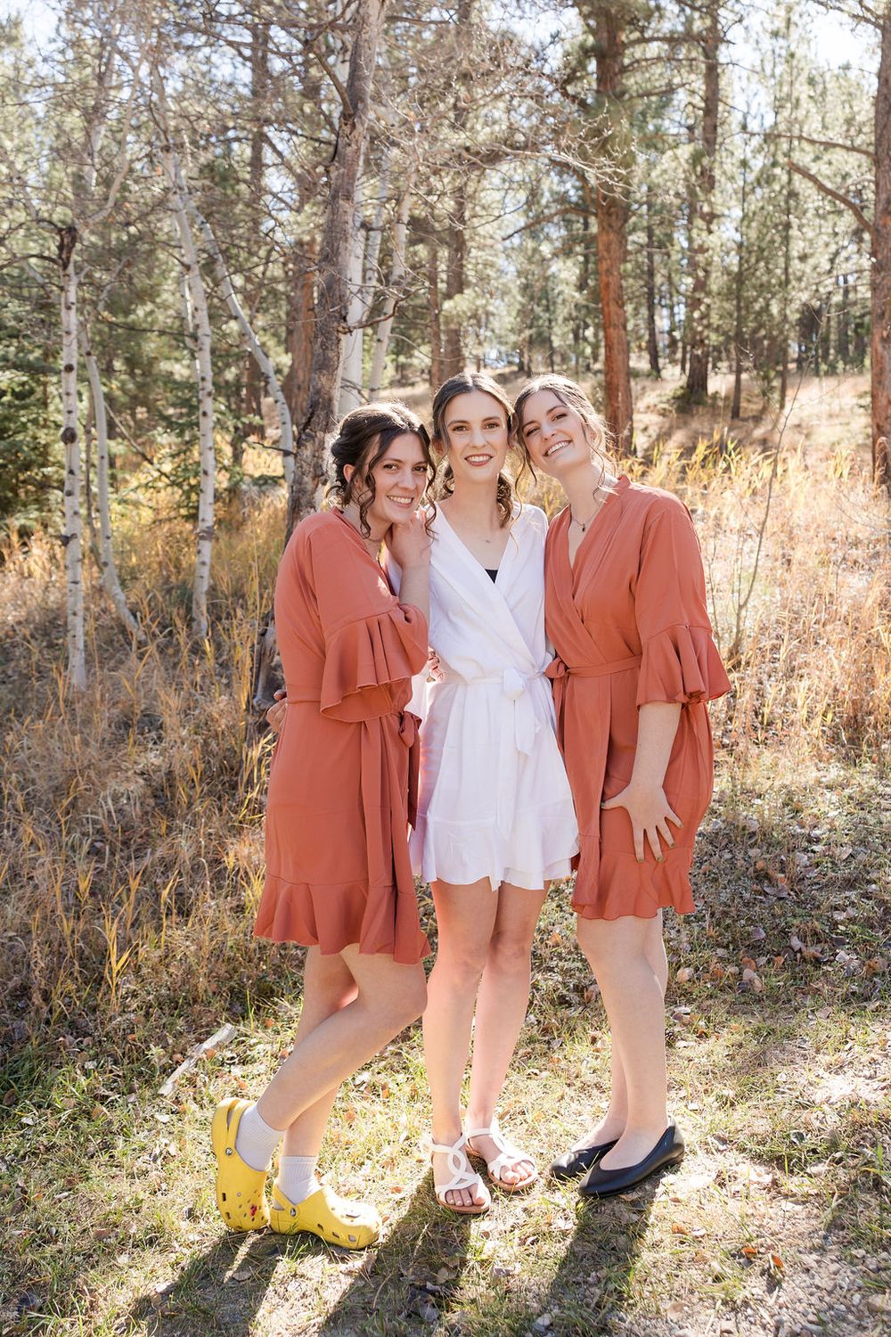 Wedding party in coral dresses poses together in a sunlit forest setting.