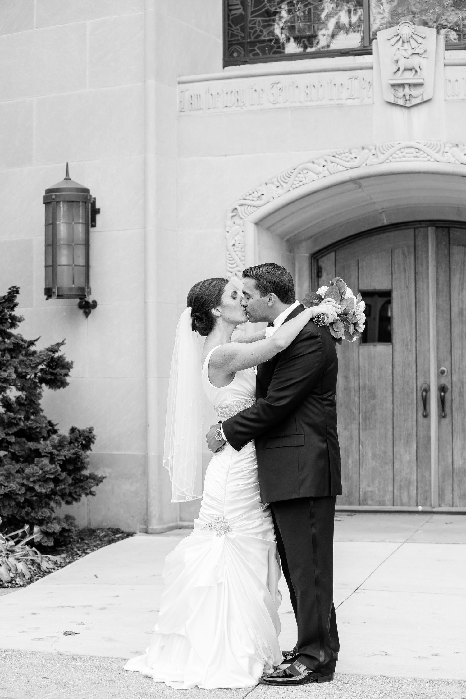 A romantic black and white wedding photo showing a couple embracing in front of an ornate church doorway.