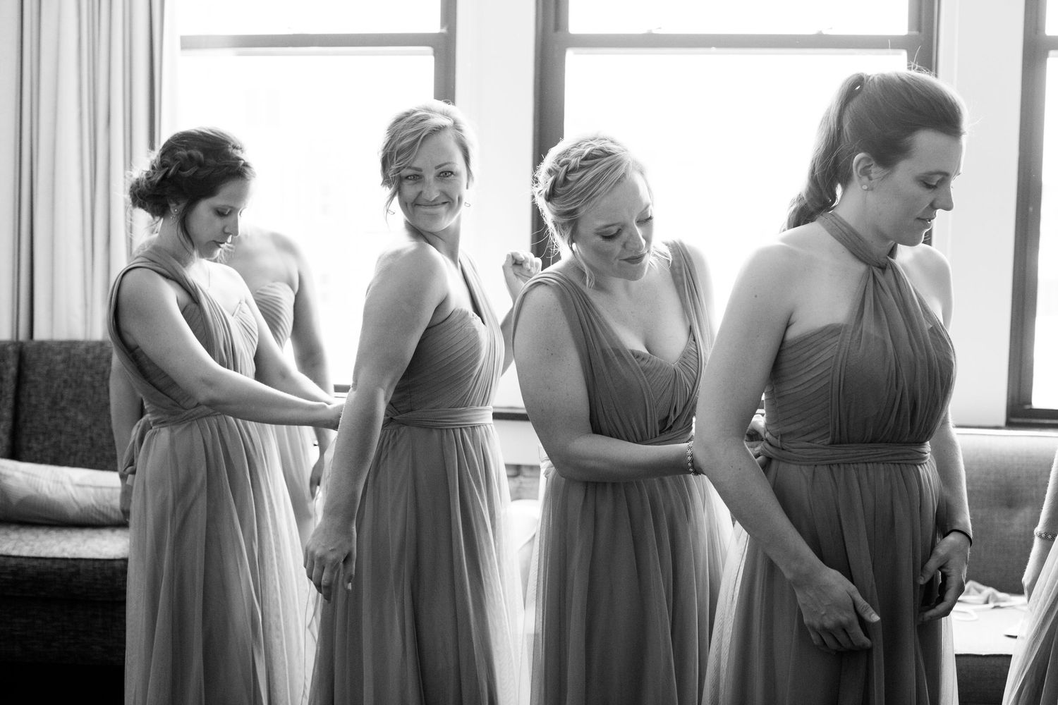Four bridesmaids in matching gray dresses get ready together in a sunlit room before a wedding ceremony.