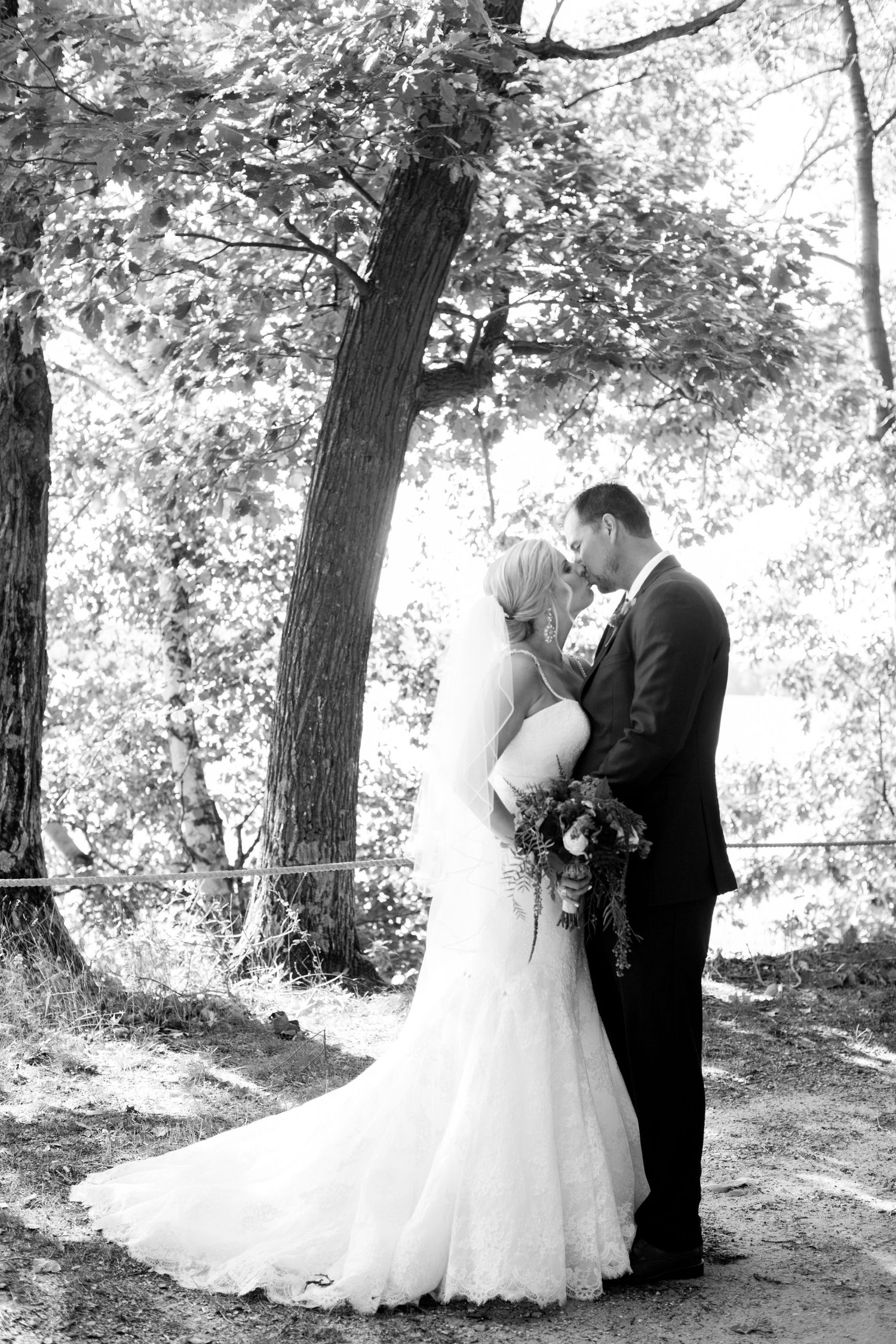 A romantic black and white wedding photo series showing a couple in formal attire embracing under trees in a natural setting.