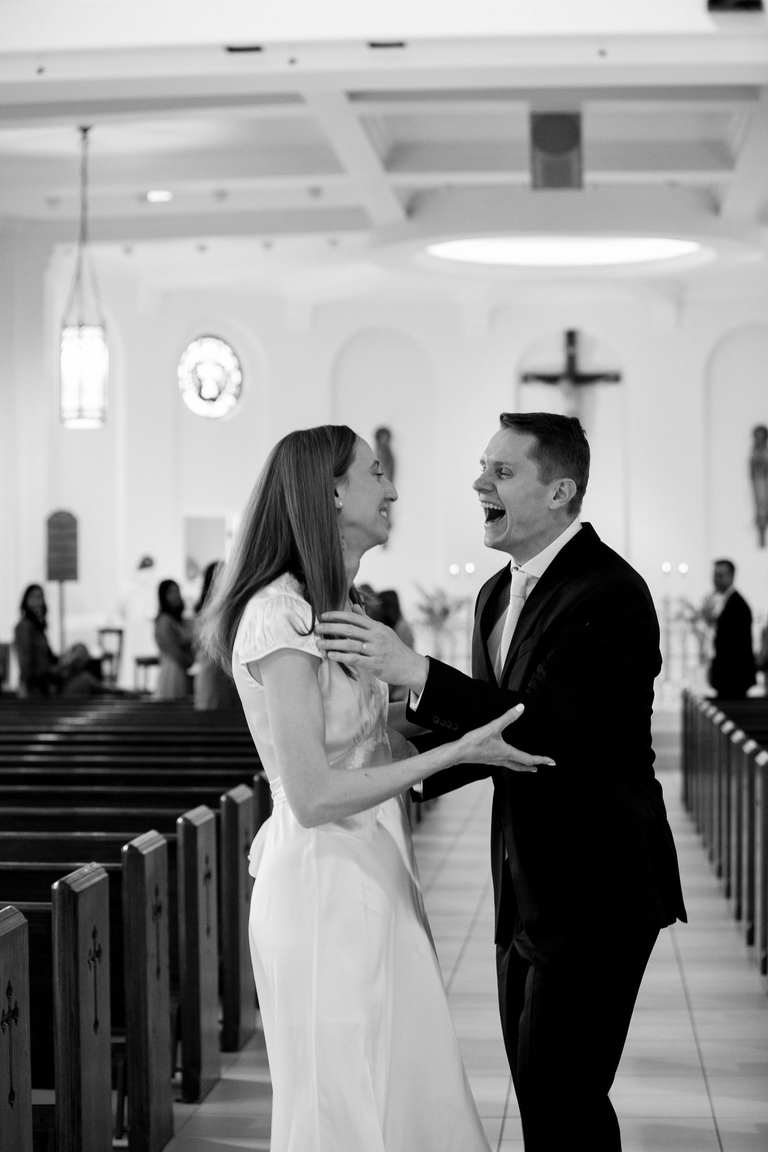 Black and white photo of a joyful bride and groom sharing a laugh in a church aisle on their wedding day.