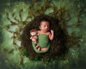 Sleeping newborn baby in green wrap holding a lion stuffed toy in a mossy nest on green backdrop.
