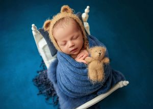 Sleeping newborn in bear ear bonnet wrapped in blue swaddle holding a small teddy bear in a white miniature bed.