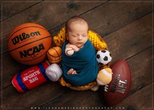 Sleeping newborn wrapped in teal swaddle nestled in a yellow bowl surrounded by sports balls including basketball and football.