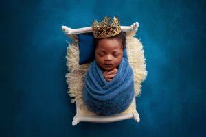Newborn baby wrapped in blue swaddle wearing a gold crown on a white mini bed.