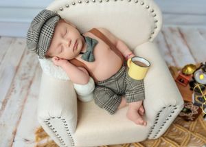 Adorable newborn in plaid newsboy cap and suspenders sleeping in a miniature armchair holding a small yellow cup.