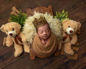 Sleeping newborn baby wrapped in brown with a gold crown flanked by two teddy bears on wood floor.