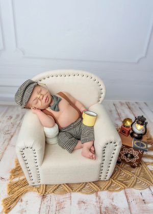 Newborn baby sleeping in a tiny armchair wearing plaid newsboy outfit holding a yellow cup with vintage props nearby.