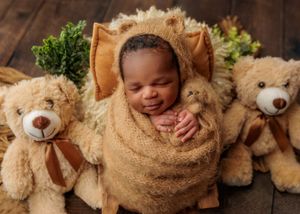 Smiling newborn wrapped in tan bear costume flanked by two teddy bears on wooden backdrop.
