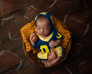Sleeping newborn baby dressed in blue and yellow football uniform holding a miniature football in a bowl.