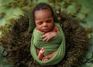 Newborn baby wrapped in green swaddle nestled in dark fur bowl with green nature backdrop.