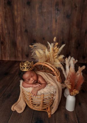 Sleeping newborn baby wearing a gold crown in a wicker basket with pampas grass decor.
