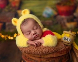 Sleeping newborn in yellow Winnie the Pooh costume resting on barrel with honey jar.