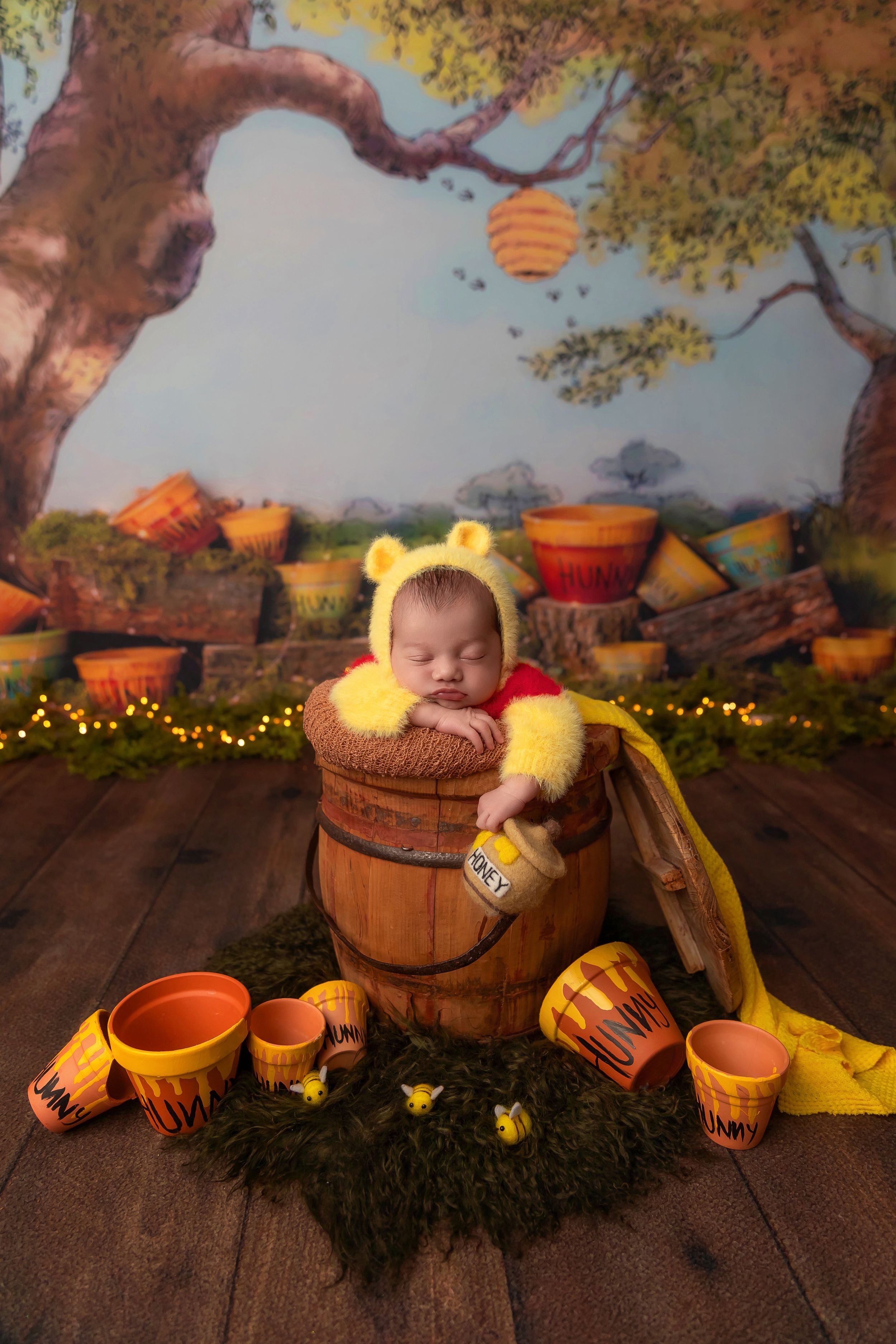 Newborn baby sleeping in a wooden honey barrel wearing Winnie the Pooh costume with yellow bear ears hat.