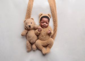 Smiling newborn in a bear outfit next to a teddy bear on a white swing.