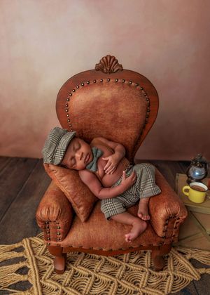 Sleeping newborn in plaid outfit curled up in a miniature brown leather armchair.