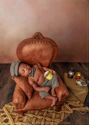 Sleeping newborn in plaid outfit posed in a miniature brown leather armchair.