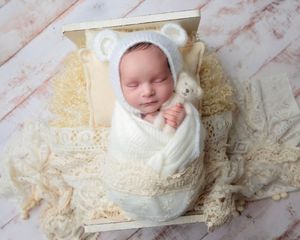 Sleeping newborn in white bear bonnet and swaddle holding tiny teddy bear.