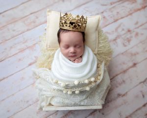 Newborn baby wrapped in white swaddle wearing gold crown on lace pillow.
