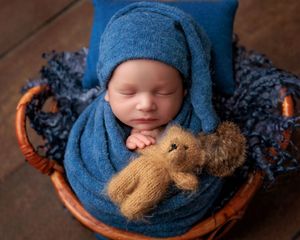 Sleeping newborn wrapped in blue knit swaddle with teddy bear in wicker basket.
