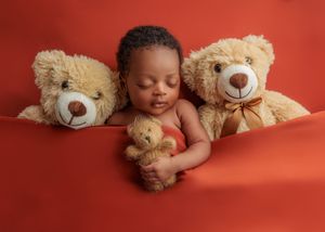 Sleeping newborn baby nestled between two large teddy bears on a red background.