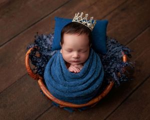 Newborn baby wrapped in blue swaddle wearing gold crown, sleeping peacefully in wicker basket with blue pillow.
