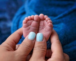 Close-up of tiny newborn baby feet held gently by adult hands with light blue nail polish on blue background.