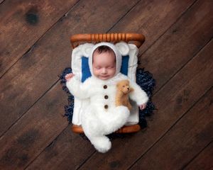 Smiling newborn in white bear onesie holding teddy bear, lying on small wooden bed on dark hardwood floor.