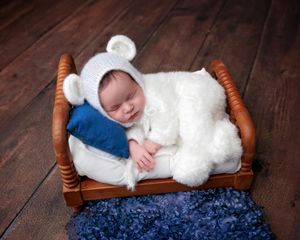 Sleeping newborn baby in white bear bonnet resting on tiny wooden bed with blue pillow on dark wood floor.