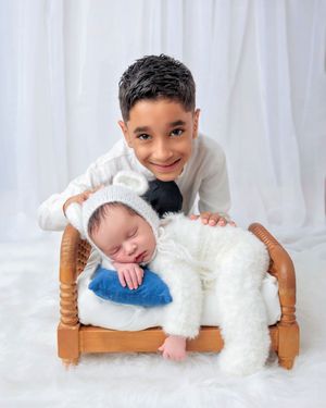 Smiling older boy proudly posing with sleeping newborn sibling in white bear outfit on small wooden bed.