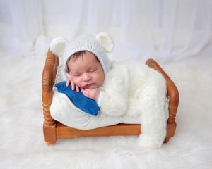 Newborn baby in white bear bonnet sleeping on tiny wooden bed with blue pillow on white fur blanket.