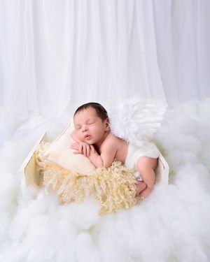 Peaceful newborn baby with white angel wings sleeping in small wooden bed surrounded by fluffy white clouds.