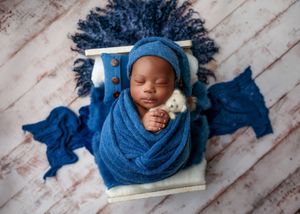 Sleeping newborn swaddled in royal blue wrap on a small white wooden bed with blue fur accents.