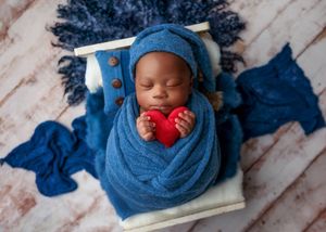 Newborn baby swaddled in blue wrap holding a red heart prop while sleeping peacefully in a wooden bed.