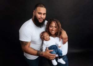 Family portrait of parents in white shirts holding their smiling baby against a dark studio background.