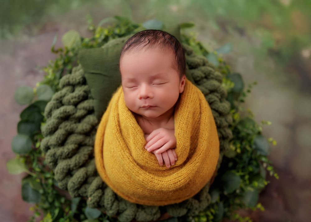 Newborn baby wrapped in golden yellow knit swaddle nestled in a green wreath with eucalyptus leaves.