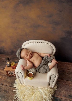 Newborn baby sleeping on miniature sofa wearing plaid outfit with tiny hat and coffee cup.