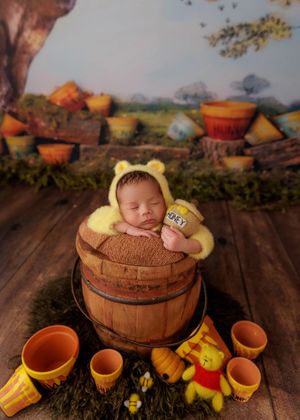 Newborn baby in Winnie the Pooh bear costume sleeping in a wooden barrel with honey pot props.