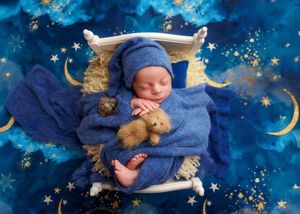Newborn baby in blue wrap and hat holding teddy bear on small white bed with starry background.