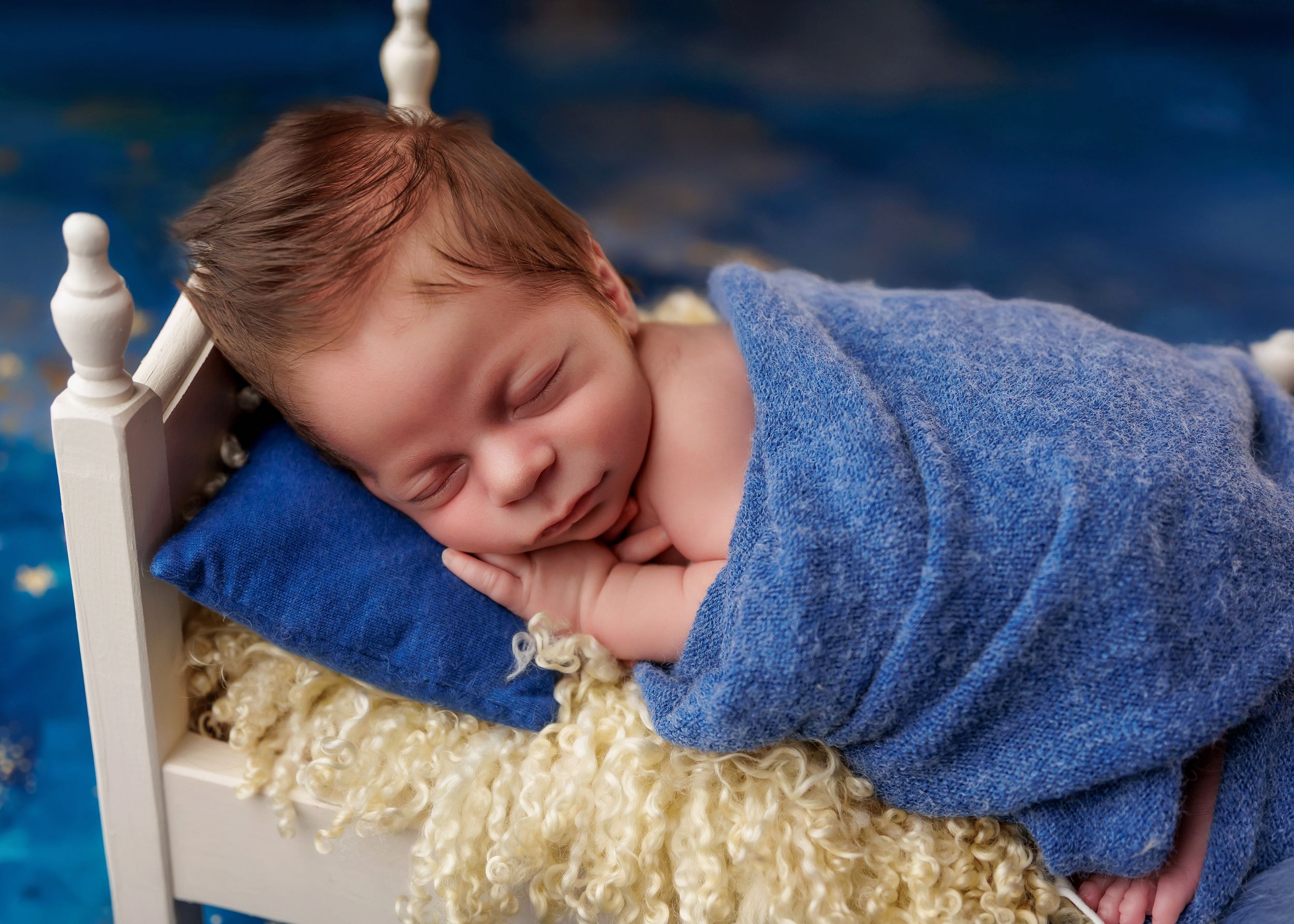 Sleeping newborn baby wrapped in blue blanket on tiny white bed with fluffy yellow bedding.