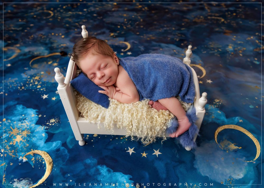 Newborn baby sleeping on miniature white bed with blue wrap on celestial moon and stars backdrop.