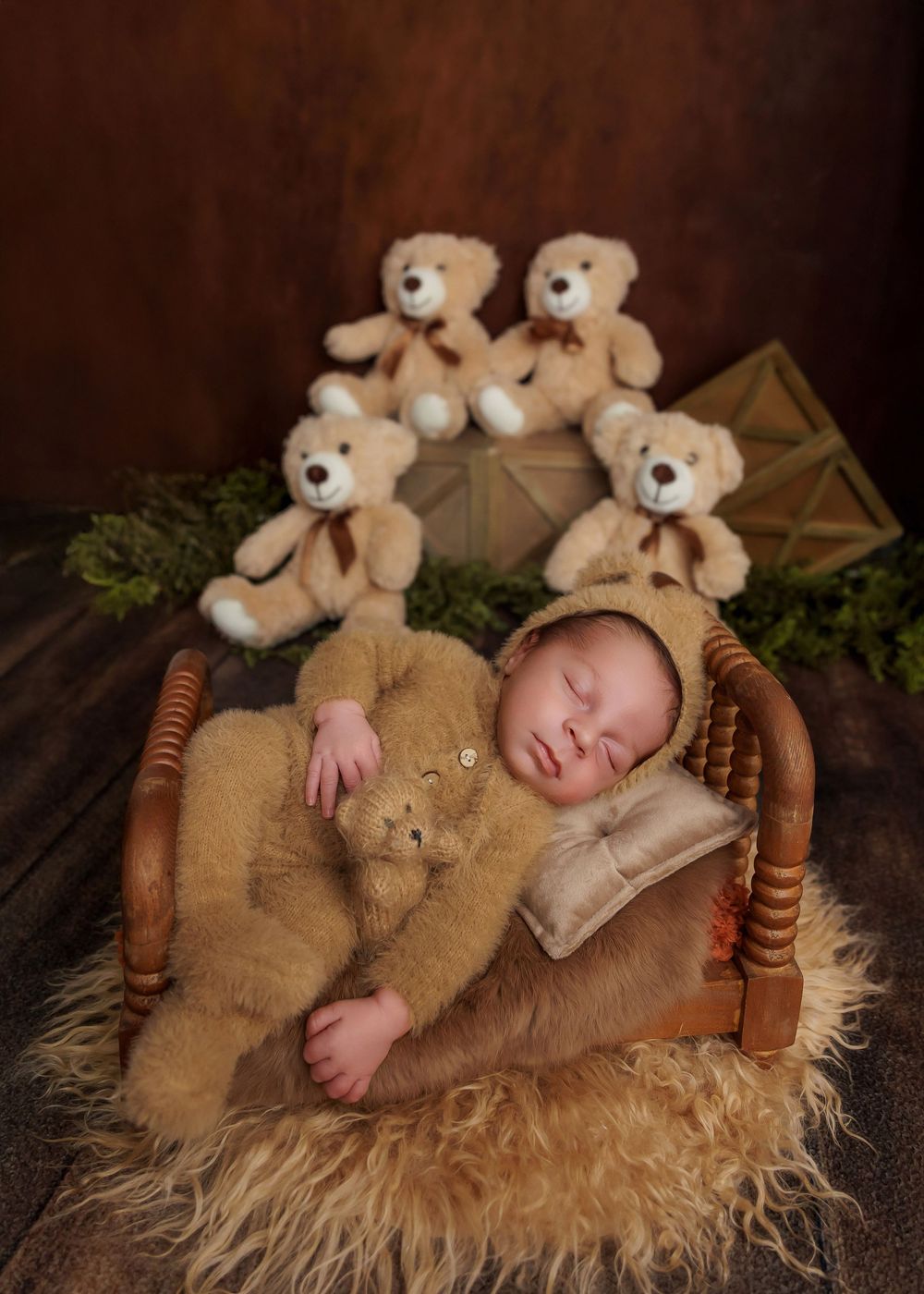 Newborn baby in bear outfit sleeping on a tiny wooden bed holding a teddy bear with plush bears behind.