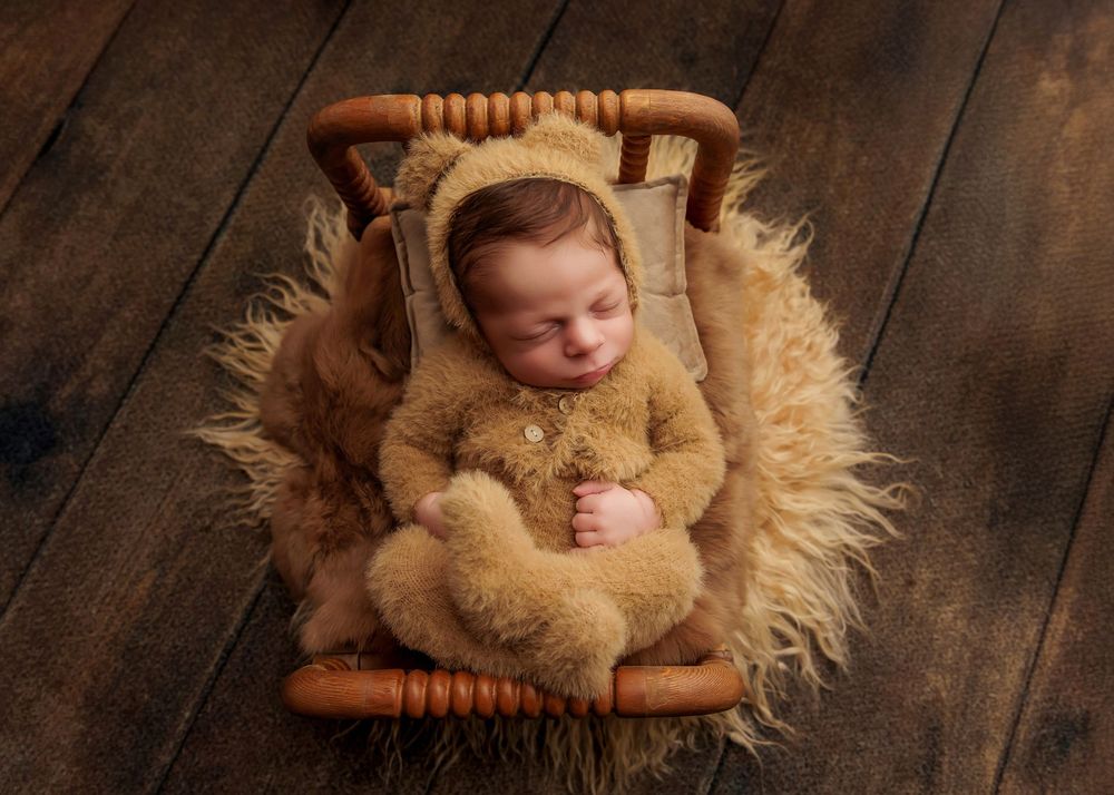 Newborn baby in brown bear romper and bonnet sleeping in a wicker basket holding a small teddy bear.