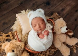 Sleeping newborn in white bear bonnet swaddled in white wrap hugging small teddy bear.