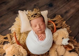 Sleeping newborn wrapped in white swaddle wearing gold crown on pillow surrounded by teddy bears.