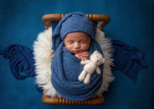 Sleeping newborn swaddled in blue wrap with matching hat holding a white teddy bear in a wooden basket.